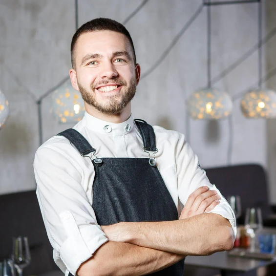 Ein Mann in Schürze mit verschränkten Armen lächelt.