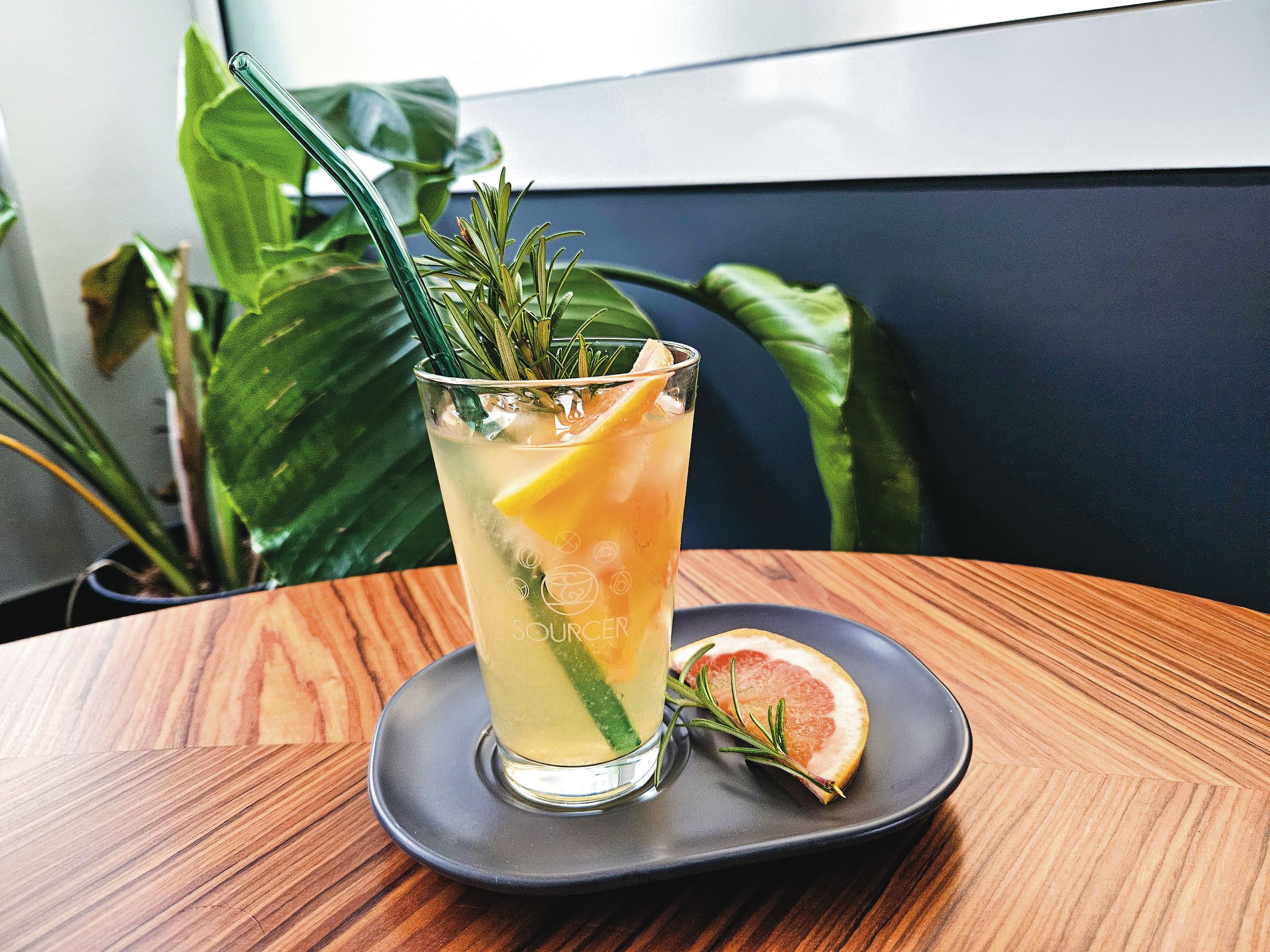 SOURCER Grüner Eistee mit Grapefruit und Rosmarin Ein Glas Getränk mit einem Strohhalm und einer Orangenscheibe auf einem Teller.